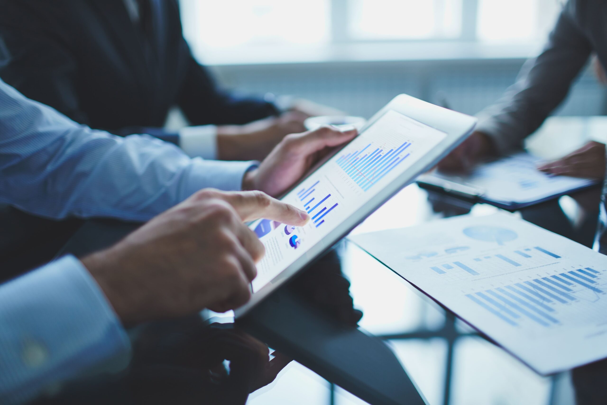 Reunião de negócios com pessoas analisando gráficos e dados em tablets e papéis, em ambiente corporativo profissional.